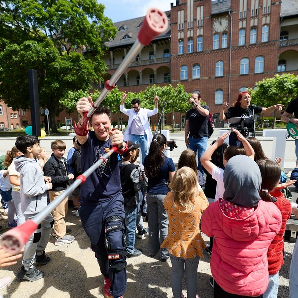 Viele Kinder stehen vor einer kleinen Bühne im Freien, auf der eine Band steht. Moderator André Gatzke hält Krücken in die Luft.