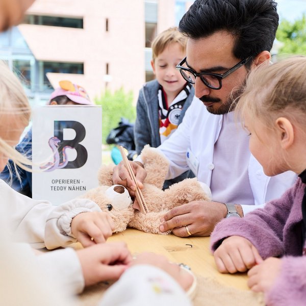 Dr. med. Nibras Naami berührt einen auf einem Tisch liegenden Teddybär mit einer Zange, fünf Kinder stehen um ihn herum.