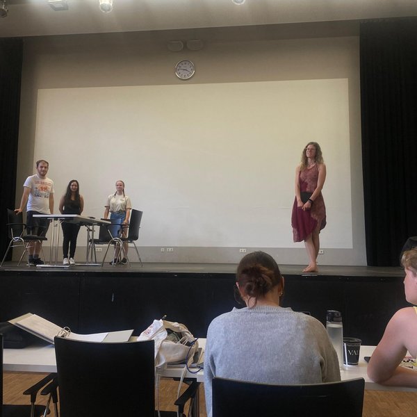 Vier Studierende stehen bei der Probe des Stückes auf der Bühne. Zwei weitere Personen sitzen an Tischen vor der Bühne.