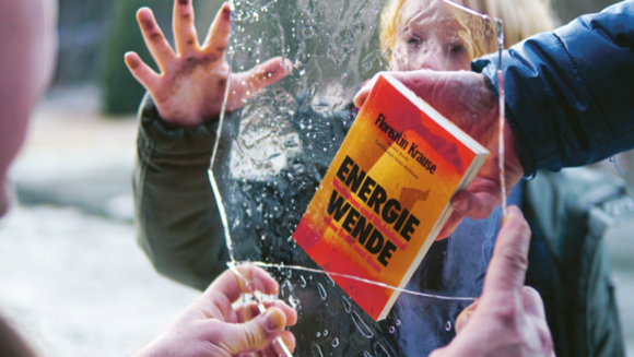 Eine Hand hält das Buch "Energiewende" gegen ein Stück einer Glasscheibe, die eine andere Person mit zwei Händen hält.