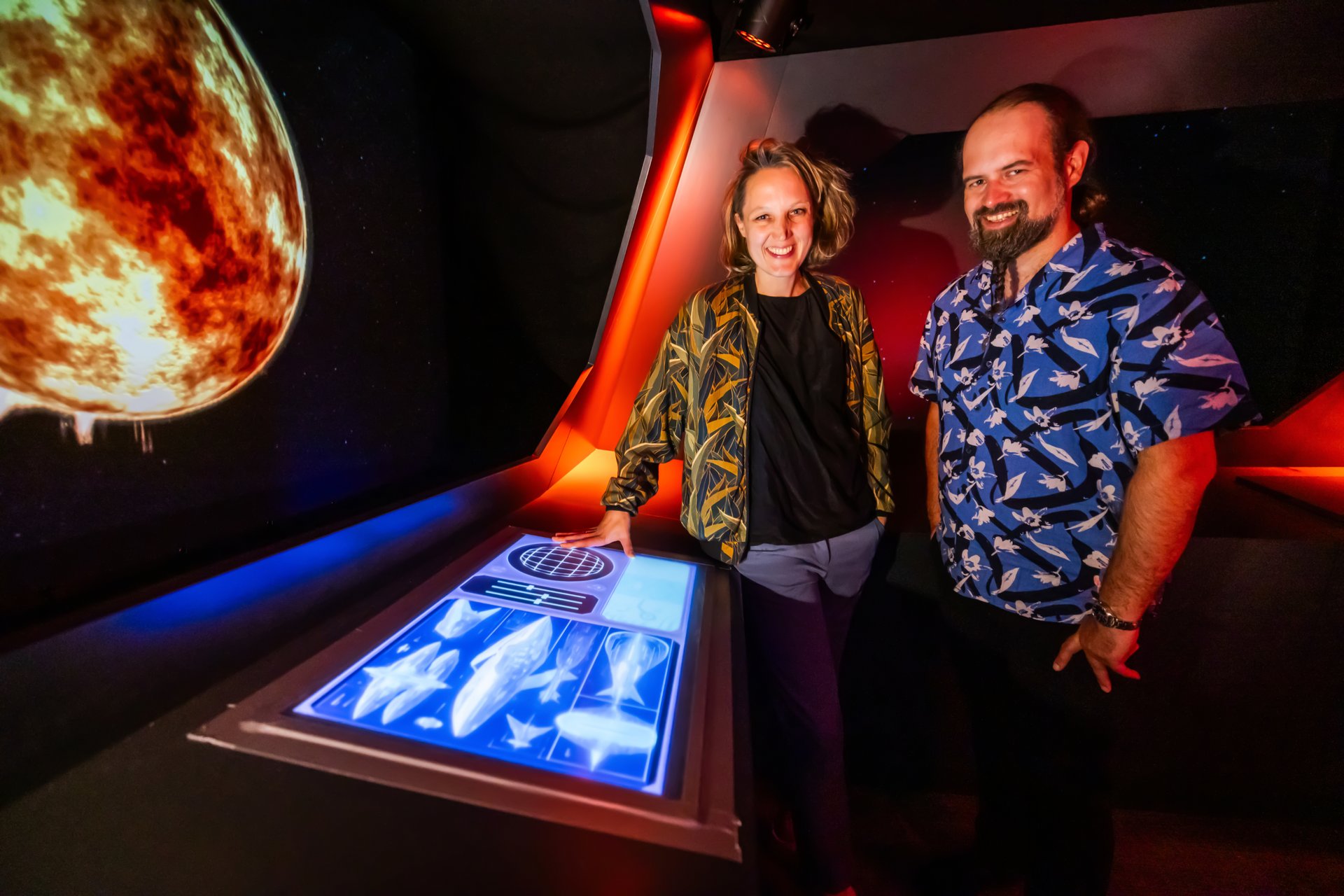 Eine Frau und ein Mann im Cockpit neben einem großen Display, der einen Planeten zeigt.
