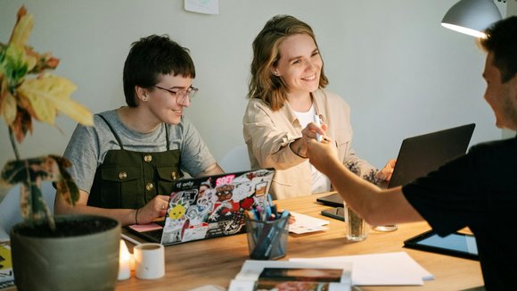 Drei Personen sitzen an einem Tisch mit Laptops, Büchern und Stiften