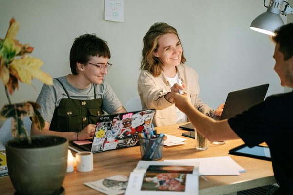 Drei Personen sitzen an einem Tisch mit Laptops, Büchern und Stiften