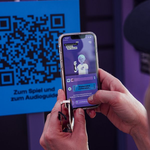 Zwei Hände halten ein Handy vor einen QR-Code, auf dem Handy ist eine illustrierte Figur und eine Skala zu sehen.