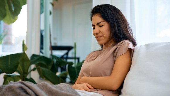 Eine junge Frau auf dem Sofa hält sich vor Schmerzen den Bauch.