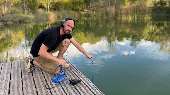 Henning Francik hockt auf einem Steg, hat Kopfhörer auf und hält ein Kabel in einen See.