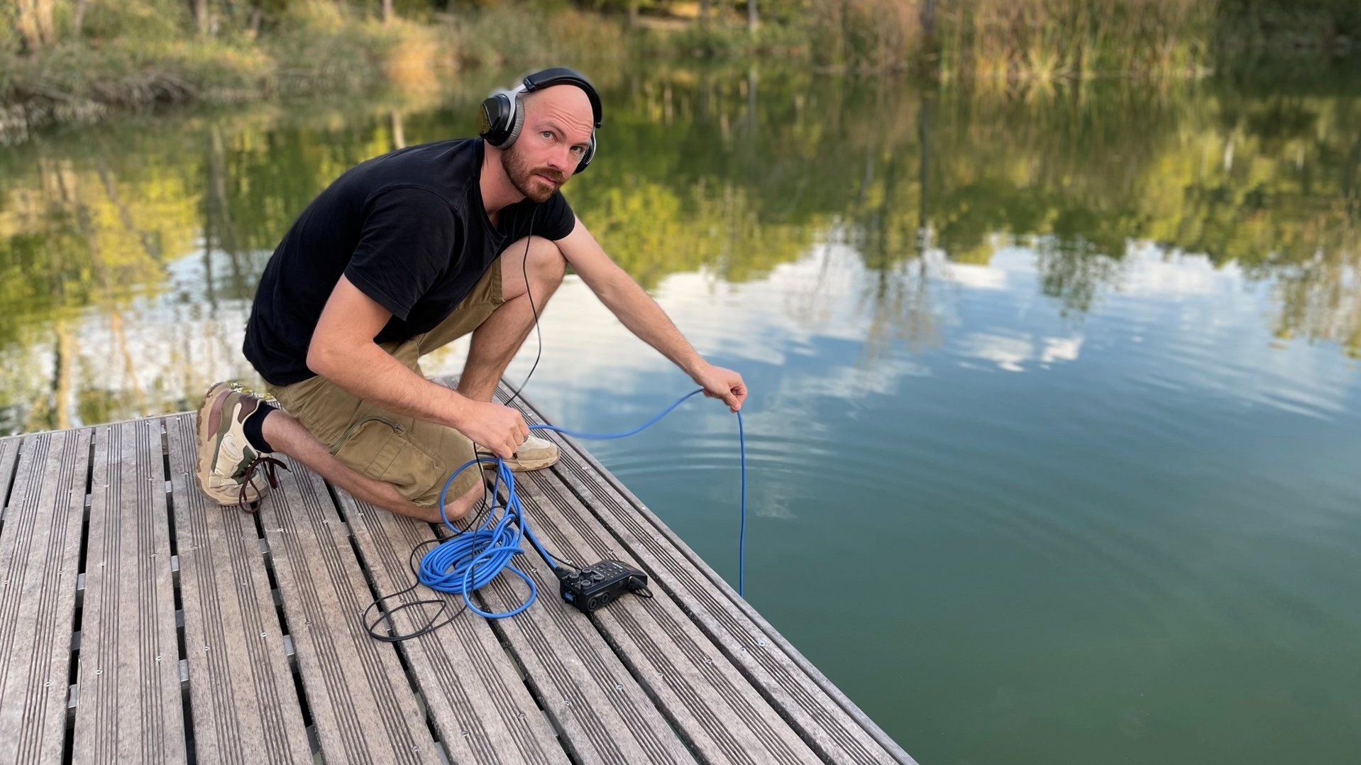 Henning Francik hockt auf einem Steg, hat Kopfhörer auf und hält ein Kabel in einen See.