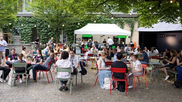 Mehrere Menschen sitzen an runden Tischen, im Hintergrund ein Zelt mit zwei Schildern mit den Aufschriften "Festival Café" und "Festival Bar"