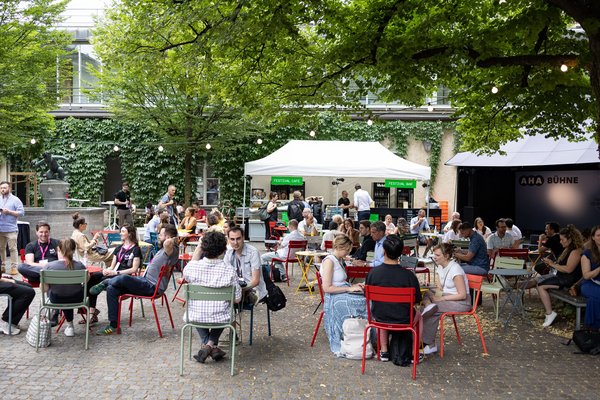 Mehrere Menschen sitzen an runden Tischen, im Hintergrund ein Zelt mit zwei Schildern mit den Aufschriften "Festival Café" und "Festival Bar"