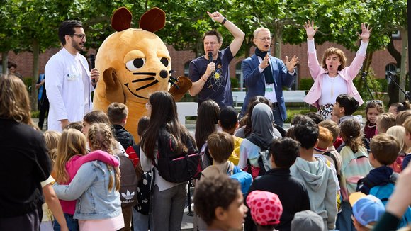 Vier Menschen und die Maus als Walking Act stehen auf einer Bühne im Freien, davor stehen viele Kinder.