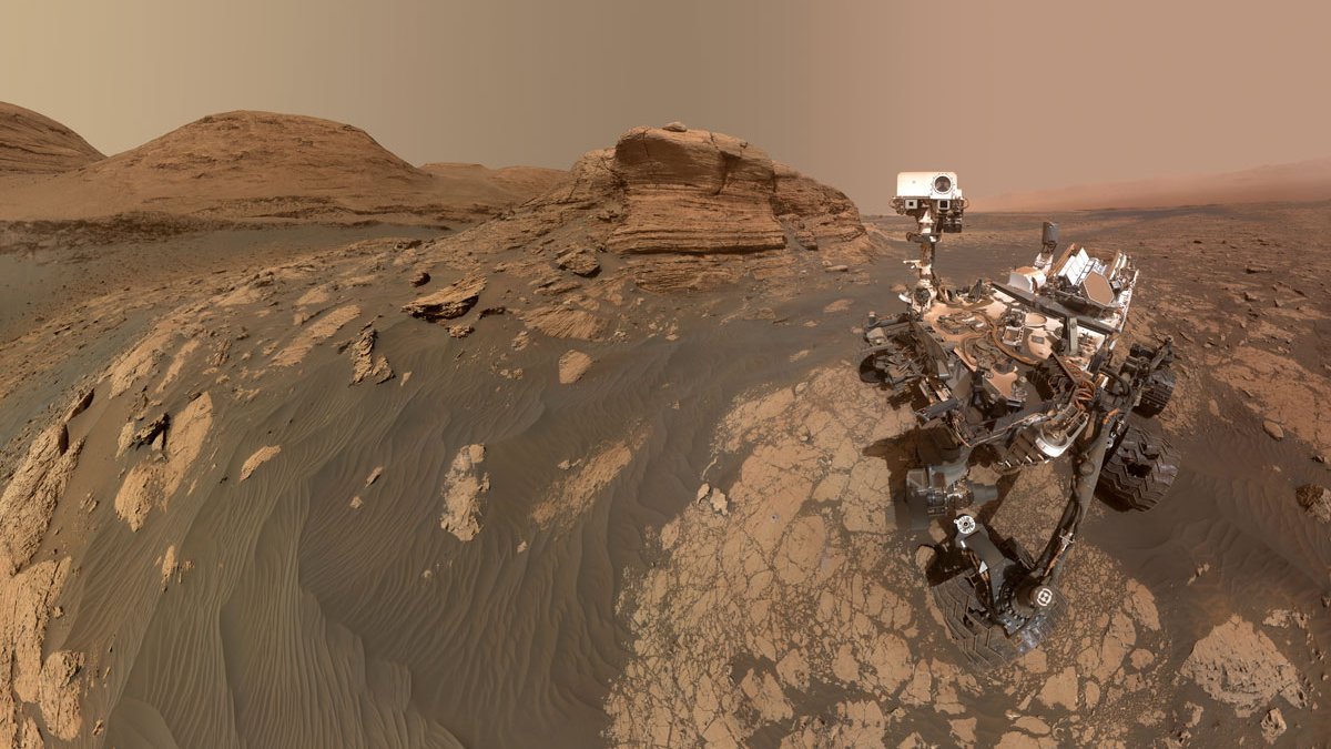 Marsrover Curiosity auf dem Mars. Marsboden und -himmel sind rötlich gefärbt. Im Hintergrund sind Berge zu sehen. 