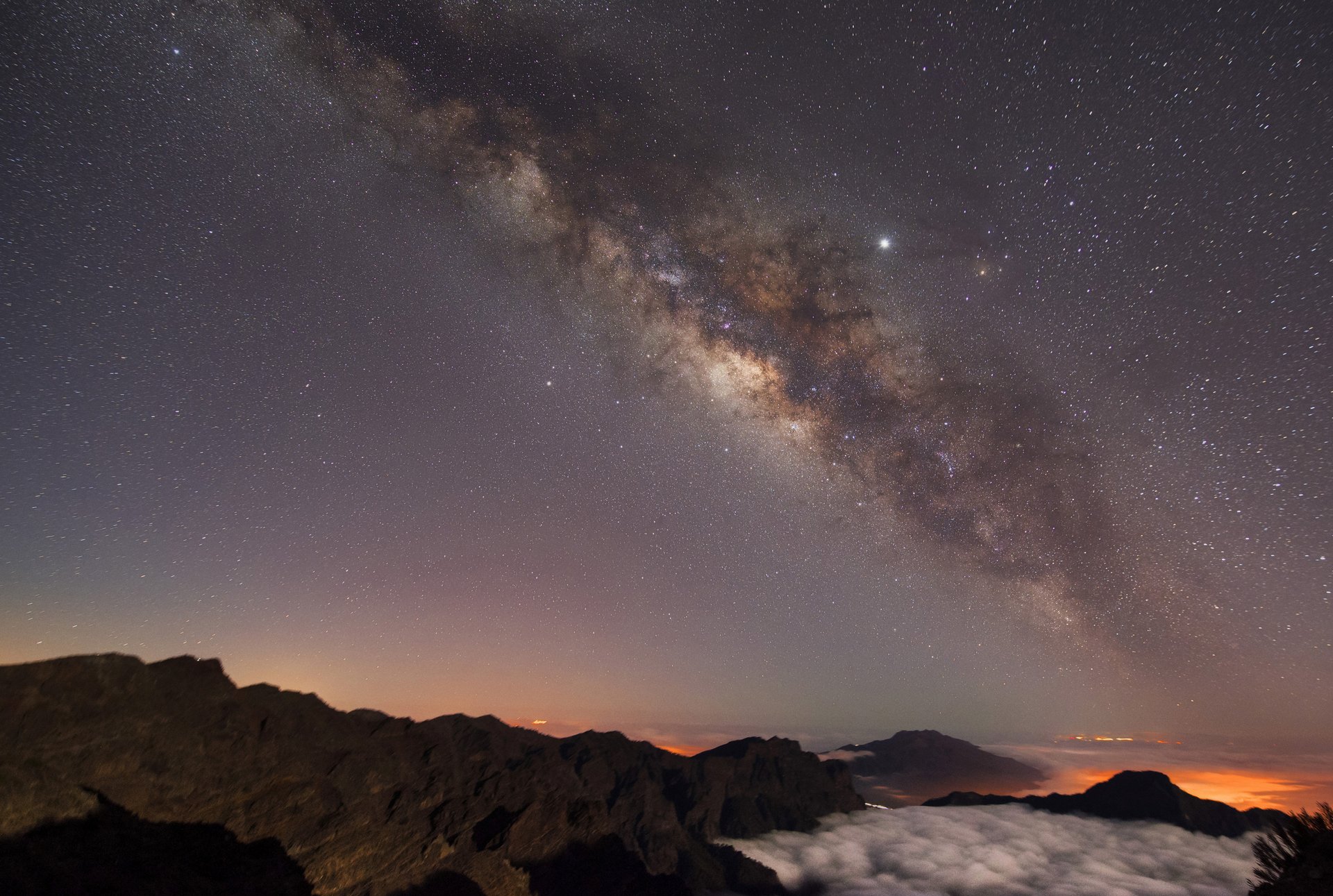 Unten ist eine dunkle Berglandschaft zu sehen, über ihr ist der Nachthimmel mit leuchtenden Sternen und einem grauen Galaxienebel.