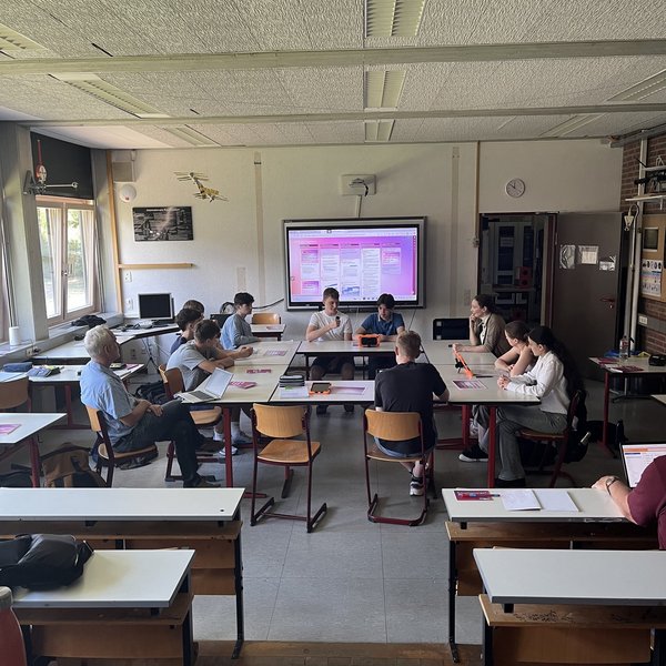 Elf Personen sitzen an zum Kreis zusammengestellten Tischen in einem Klassenraum.