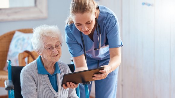 Frau in Pflege-Kleidung steht neben einer sitzenden, älteren Frau und hält ihr ein Tablet hin.