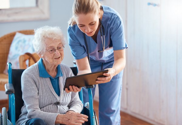 Frau in Pflege-Kleidung steht neben einer sitzenden, älteren Frau und hält ihr ein Tablet hin.