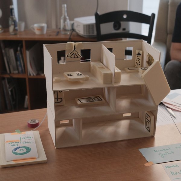 Ein kleines, gebasteltes Modellhaus befindet sich auf einem Tisch. Durch eine fehlende Hauswand zeigt sich die Raumaufteilung und Einrichtung des Hauses.