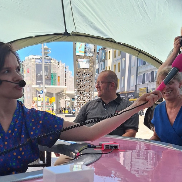 Eine Frau sitzt in der Mitte eines runden Tisches und hält ein Mikrofon zum Tischrand.