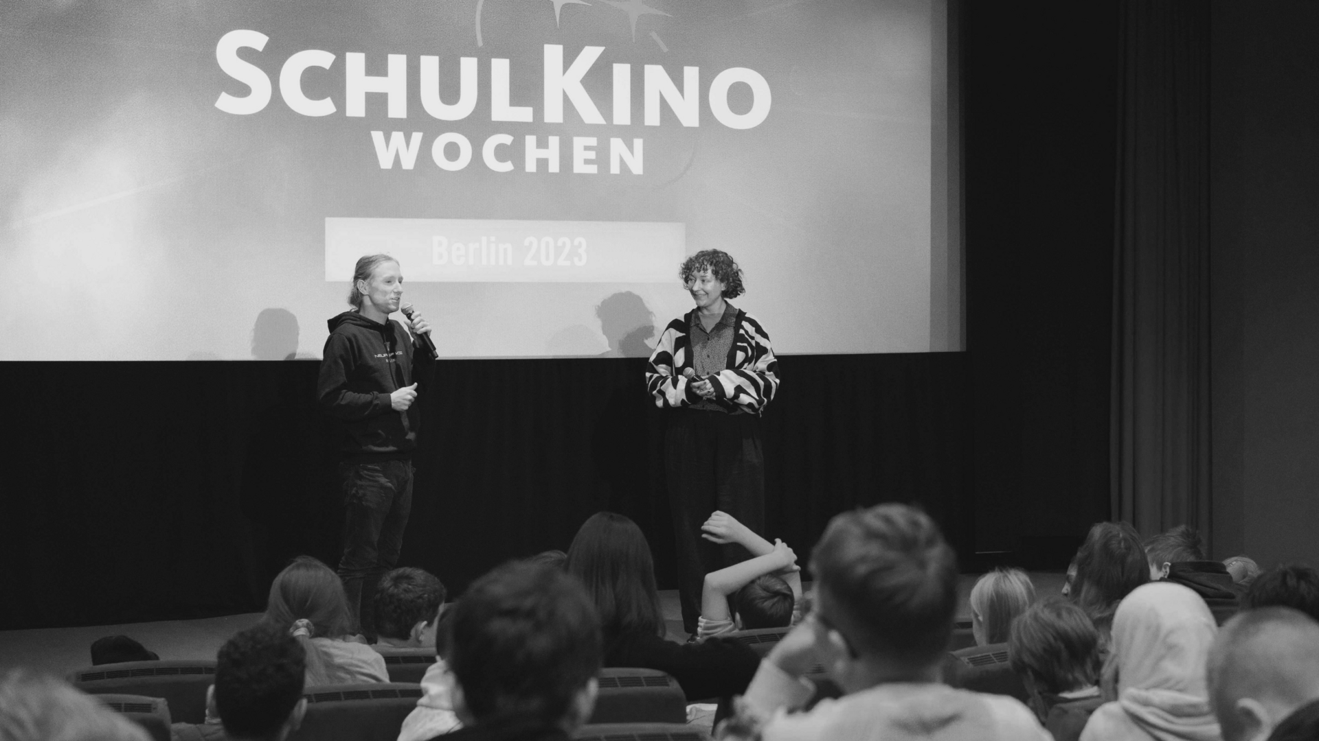 Projektbild SchulKinoWochen: Foto von zwei jungen Erwachsenen, die beide ein Mikrofon halten. Sie stehen vor einer Gruppe Schülerinnen und Schüler in einem Kinosaal und unterhalten sich.