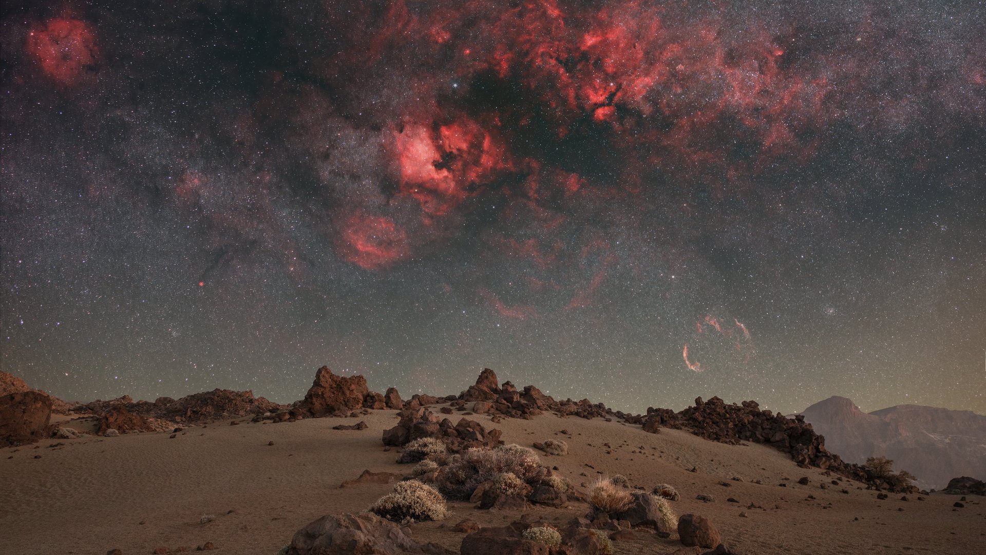 Unten ist eine steinige Wüste zu sehen. Der Himmel ist voller leuchtender Sterne und einem dunkelroten Galaxienebel.