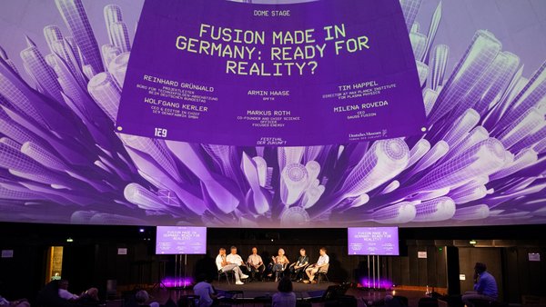 Sechs Personen sitzen auf einer Bühne vor einem Publikum, auf dem Kuppeldach darüber ist auf einem Screen der Panel-Titel “Fusion Made in Germany: Ready for Reality?” mit den Namen der Diskutierenden eingeblendet.  