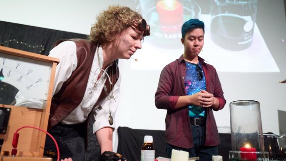 Verkleidete*r Schauspielerin und Schauspieler führen ein Live-Experiment durch. Sie stülpen ein großes Glas über eine Kerze. 