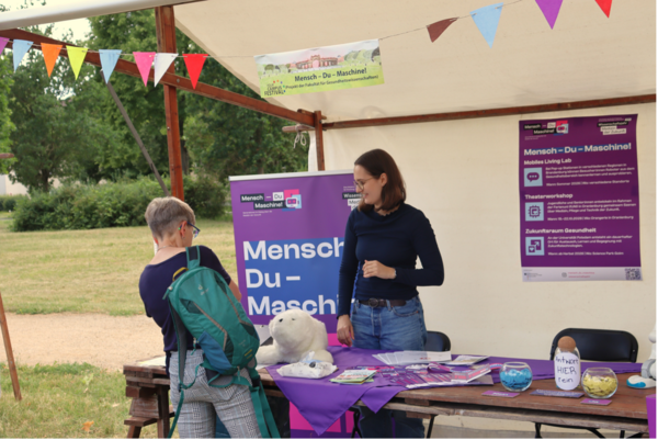 Informationsstand des Projekts Mensch – Du – Maschine! Projektmitarbeiterin ist im Austausch mit sich informierender Person.