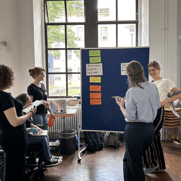 Eine Gruppe steht vor einer Pinnwand mit Zetteln und diskutiert.