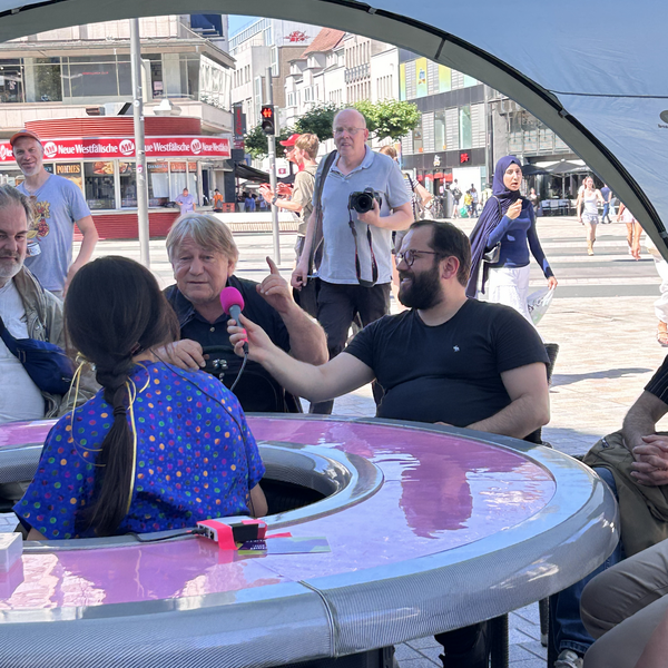 Ein Mann sitzt mit anderen am runden Tisch und spricht in ein Mikrofon.