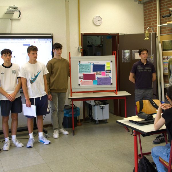 Sechs junge Personen stehen vorne im Klassenraum, an den Tischen im Raum sitzen weitere Personen.