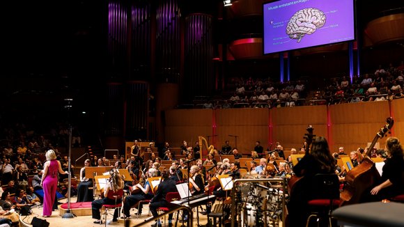 Konzertsaal mit Orchester, im Hintergrund Leinwand mit Illustration des Gehirns und dem Text: Musik entsteht im Kopf!