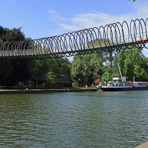 Fußgängerbrücke führt über einen Fluss, dahinter liegt ein Schiff mit Deutschlandfahne am gegenüberliegenden Ufer an einem Ankerplatz