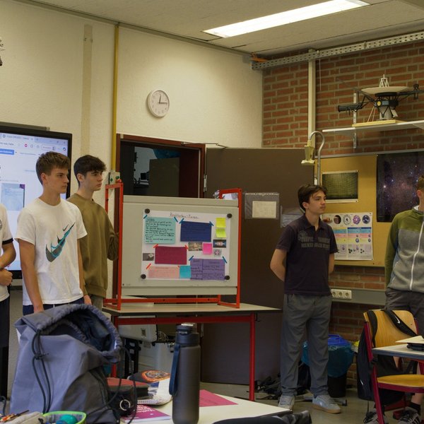 Sechs junge Personen stehen vorne im Klassenraum, an den Tischen im Raum sitzen weitere Personen.