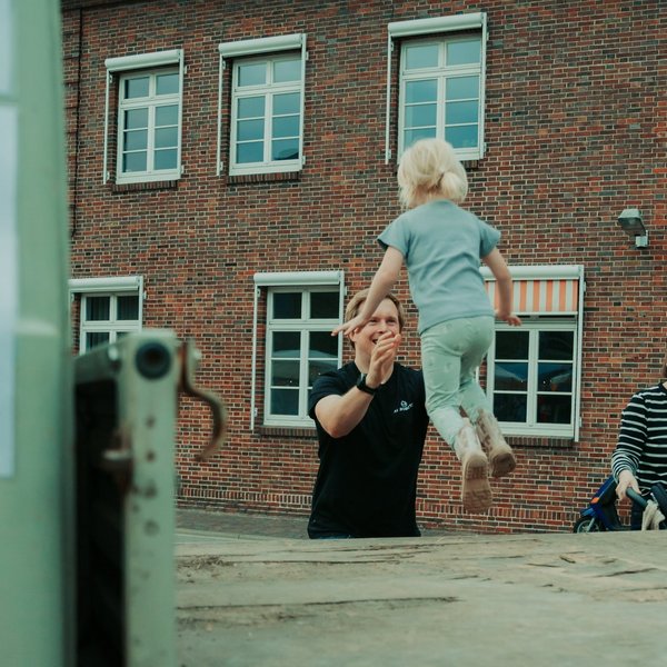 Kind springt von der Ladefläche eines Autos in die Arme eines Mannes, daneben steht eine Frau und grinst.