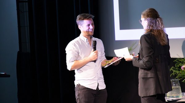Patrick Niedermeyer steht auf einer Bühne, hält ein Mikrofon in der Hand und nimmt lächelnd eine Urkunde von einer Frau entgegen