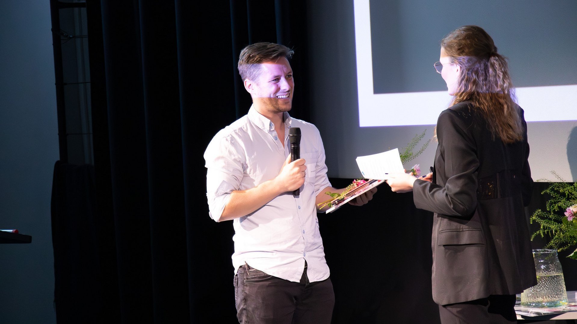 Patrick Niedermeyer steht auf einer Bühne, hält ein Mikrofon in der Hand und nimmt lächelnd eine Urkunde von einer Frau entgegen