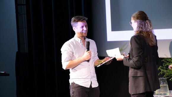 Patrick Niedermeyer steht auf einer Bühne, hält ein Mikrofon in der Hand und nimmt lächelnd eine Urkunde von einer Frau entgegen