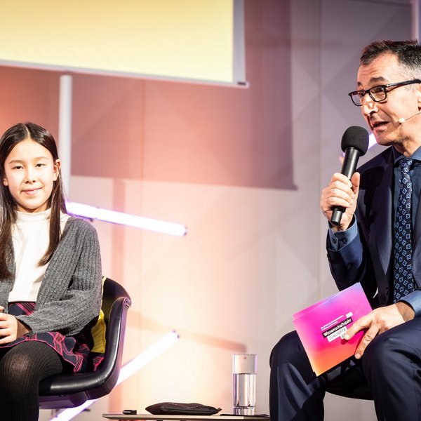 Bundesminister für Bildung und Forschung Cem Özdemir sitzt neben einem Mädchen und spricht in ein Mikrofon.
