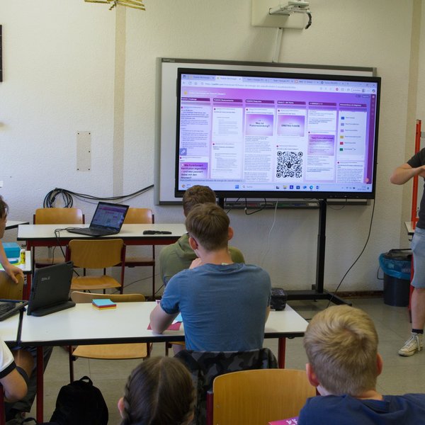 Person steht vor sitzenden Menschen in einem Klassenraum, im Hintergrund eine Präsentation.