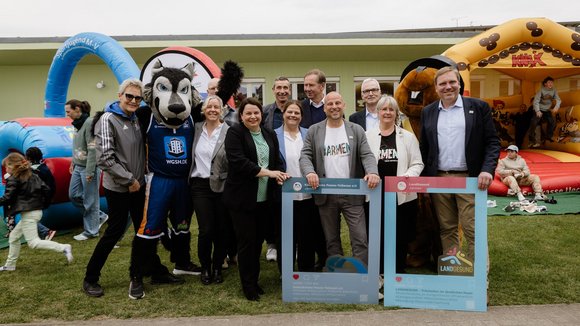 Gruppenfoto im Freien bei Auftaktveranstaltung. Mehrere Personen stehen lächelnd neben einem Maskottchen der Rostocker Seawolves.