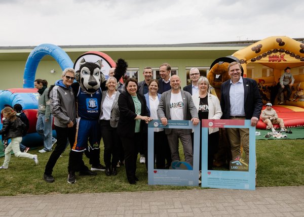 Gruppenfoto im Freien bei Auftaktveranstaltung. Mehrere Personen stehen lächelnd neben einem Maskottchen der Rostocker Seawolves.
