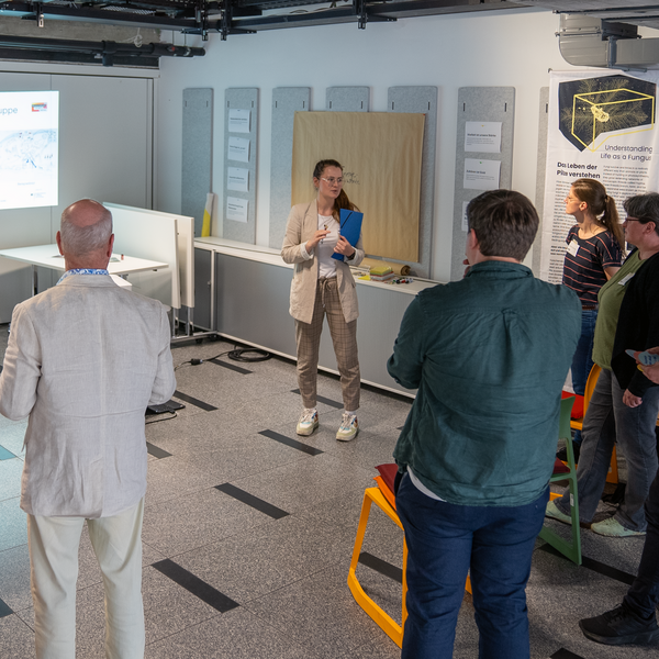 Eine Person hält einen Power-Point-Vortrag. Um sie herum stehen mehrere Personen in einem losen Halbkreis.