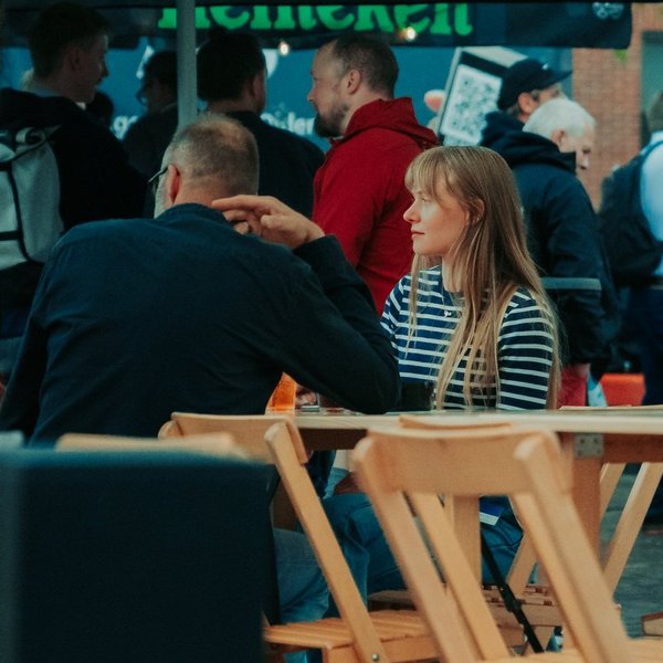 Drei Personen sitzen an Tischen auf einem Straßenfest und unterhalten sich, im Hintergrund stehen weitere Personen.