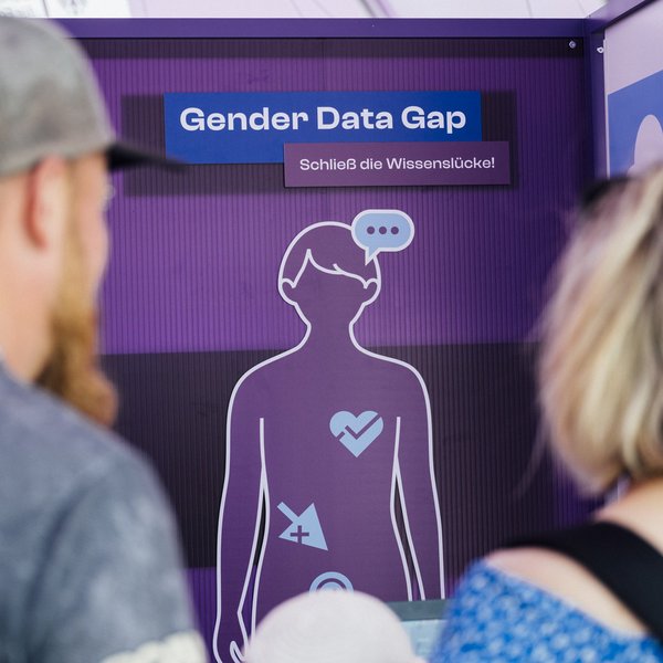Zwei Personen stehen vor einer Tafel mit dem Umriss einer Figur und dem Text "Gender Data Gap – Schließe die Wissenslücke".