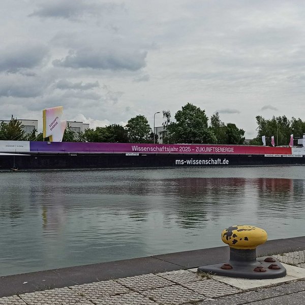 Schiff mit Aufschrift "Wissenschaftsjahr 2025 – ZUKUNFTSENERGIE" liegt am gegenüberliegenden Ufer an Ankerplatz