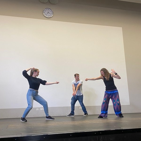 Drei Studierende stellen spielerisch auf der Bühne eine Schwert-Kampfszene nach. 