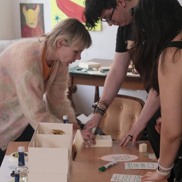 Drei Personen gemischten Alters stehen über einen Tisch gebeugt. Auf dem Tisch befinden sich ein Modellhaus und verschiedene Schreibutensilien.