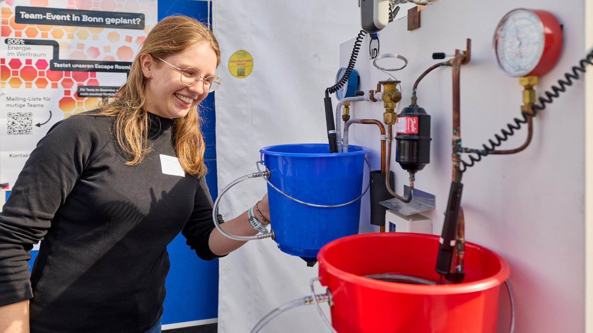 Eine Frau steht vor einer Wand mit Schläuchen, Ventilen und zwei Wassereimern.