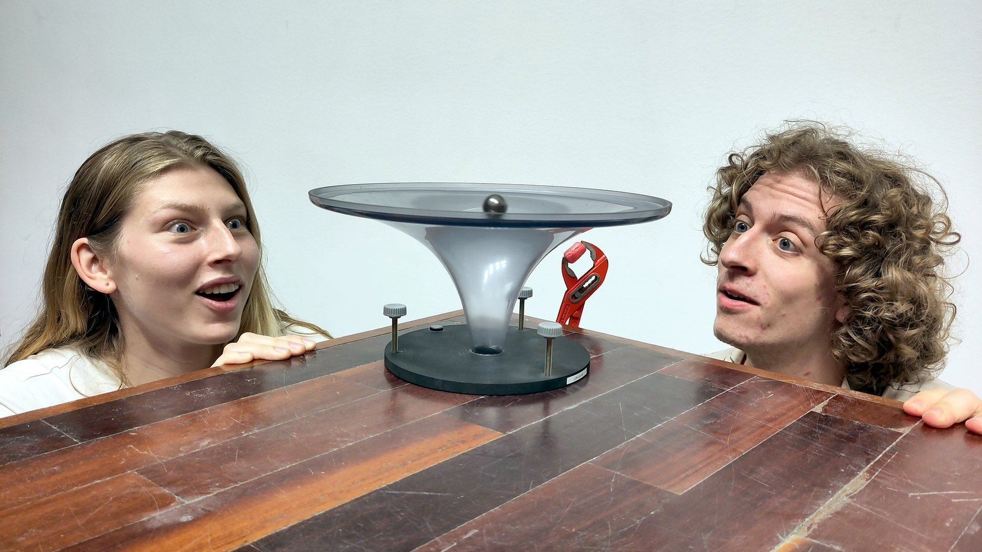 Zwei junge Forschende staunen über eine Apparatur auf einem Holztisch.