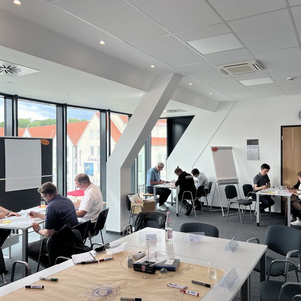 Es sitzen mehrere Kleingruppen an einzelnen Tischen und arbeiten konzentriert. Sie befinden sich in einem großen Büro.