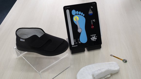 Ein Hausschuh steht neben einer Tafel, auf der ein Fuß gemeinsam mit Angaben zum Druck, zur Feuchtigkeit und zur Temperatur abgebildet ist